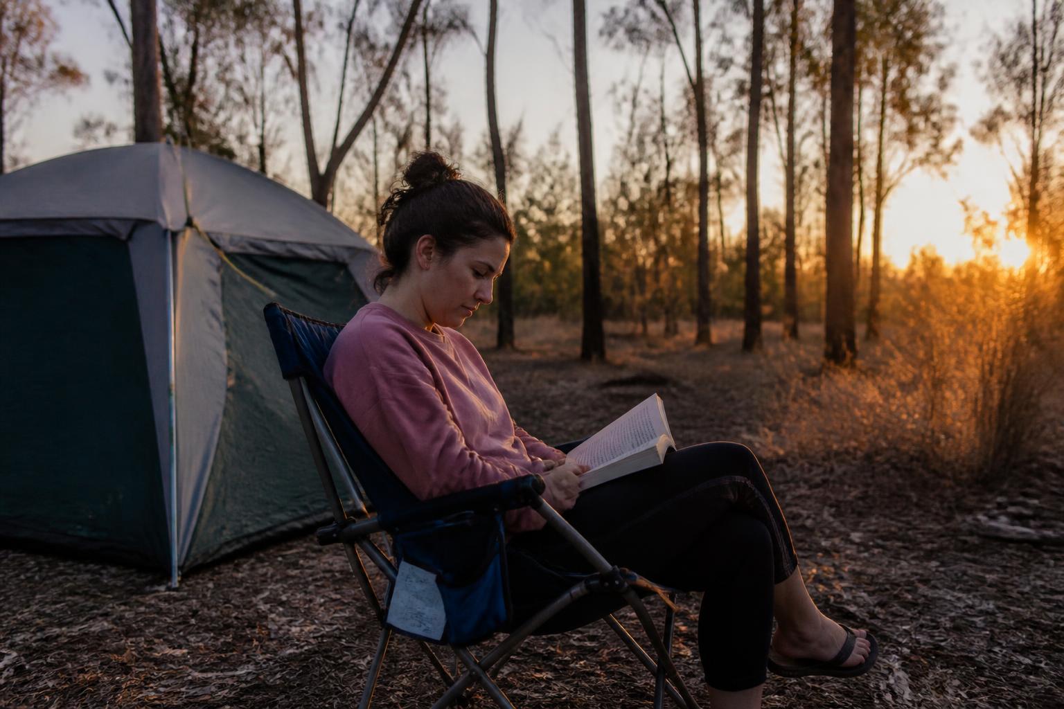 A solo camper reading in a camp chair beside her tent at golden hour